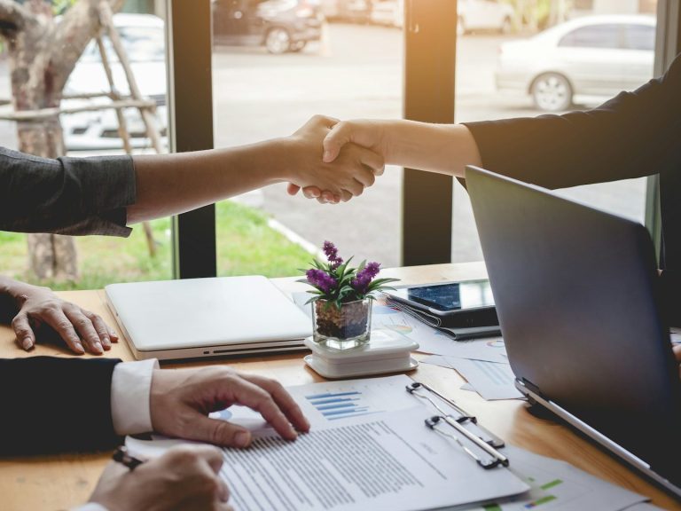 Deux personnes se serrent la main, documents en arrière-plan, sur une table. Deux personnes se serrent la main, documents en arrière-plan, sur une table.